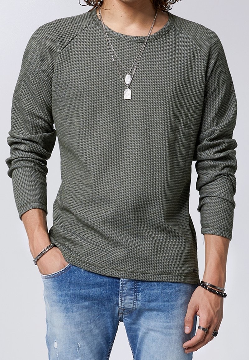 Chemise en maille texturée vert olive à manches longues raglan, associée à un denim bleu clair déchiré, et superposée avec des colliers en argent.
