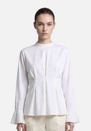 Giovane donna con i capelli tirati indietro che indossa una camicetta bianca a maniche lunghe con polsini svasati e pantaloni color crema su uno sfondo uniforme.