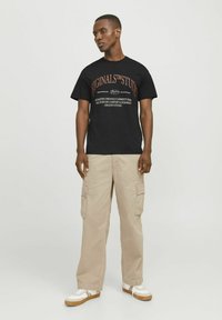 Zwarte katoenen t-shirt met rode en witte tekst, gecombineerd met lichtbeige cargobroek met zijzakken, en witte sneakers met tankleurige zolen.