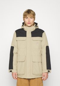 Kaki vanntett parka-jakke med svarte detaljer, med glidelås foran, justerbar hette og flere frontlommer.