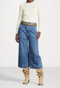 Person klädd i en krämfärgad kabelstickad tröja, vida blå jeans med dekorativt bälte och tanfärgade, slappa stövlar som står mot en vit bakgrund.