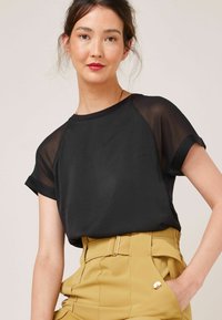 Jeune femme aux cheveux foncés portant un haut noir à manches transparentes et un pantalon moutarde taille haute, main dans la poche, fond neutre.