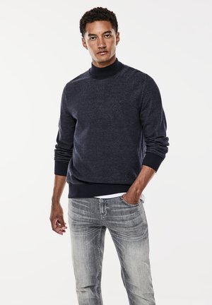 Marineblå pullover-sweater med høj hals, ribbede manchetter og kant. Bæres med lysegrå jeans med et falmet mønster. Glat, strikket tekstur.