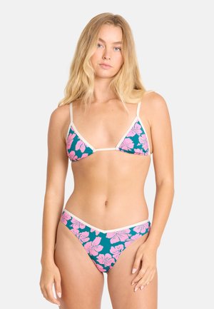 Frau mit langen blonden Haaren, die einen türkisfarbenen Bikini mit pinkem Blumenmuster und weißem Rand trägt, steht vor einem schlichten hellen Hintergrund.