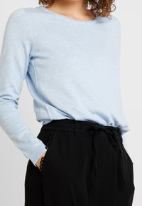 Maglione azzurro chiaro a maniche lunghe con scollatura rotonda, tessuto testurizzato, abbinato a pantaloni neri a vita alta con cintura annodabile e rifiniture decorative.