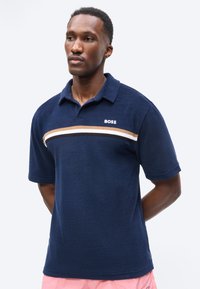 Άνδρας που φοράει μπλε σκούρο πόλο με ρίγες σε λευκό, μπεζ και μαύρο και το κείμενο «BOSS», συνδυασμένο με ανοιχτό ροζ σορτς, στέκεται με τα χέρια πίσω από την πλάτη.