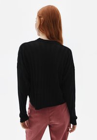 Schwarzer, gerippter Strickpullover mit langen Ärmeln und lockerem Schnitt, über rosa Hosen getragen. Das Design zeichnet sich durch vertikale Linien für Struktur aus.