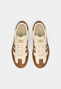 Μπεζ και καφέ δερμάτινα αθλητικά παπούτσια με τρεις καφέ ρίγες, κλείσιμο με κορδόνια και χρυσό λογότυπο στη γλώσσα. Υφή σόλας από καουτσούκ.