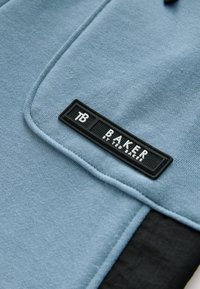 Niebieska tkanina z czarną prostokątną łatą z logo Ted Baker zszytą blisko złożonego brzegu.