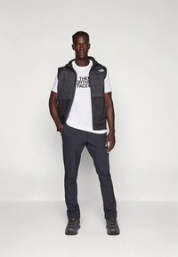 Čierna prešívaná vesta cez biele tričko s logom, doplnená tmavými nohavicami a čiernymi turistickými topánkami; hladké textúry a ležérny dizajn.