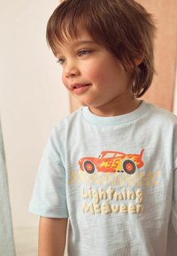 Šviesiai mėlynas trumpomis rankovėmis marškinėliai su raudonos lenktyninio automobilio grafika ir „Lightning McQueen“ tekstu smėlio spalvos, langelių raštu po automobiliu.
