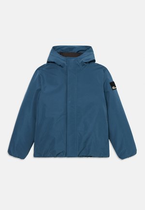 Giacca impermeabile blu con cappuccio, polsini elasticizzati, taglio dritto e una piccola etichetta con logo sulla manica. Texture liscia, design minimalista.