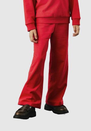Enfant portant un haut à manches longues rouge et un pantalon, avec des chaussures noires à semelle épaisse, se tenant devant un fond uni.