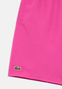 Pantalones cortos rosas hechos de tela ligera, con un pequeño logo de cocodrilo verde bordado en la esquina inferior izquierda y un bolsillo lateral.