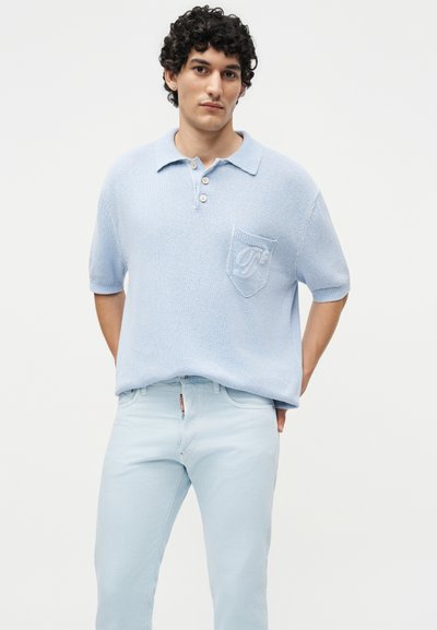 Νέος άνδρας με σγουρά μαλλιά που φοράει ανοιχτό μπλε πλεκτό πόλο με κοντά μανίκια και ανοιχτό μπλε τζιν, στέκεται μπροστά σε ένα απλό φόντο.