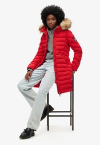 Rød puffjakke med en faux fur hætte, lynlås lukning og sidelommer, båret med en grå sweater og lyseblå jeans, siddende på en sort skammel.