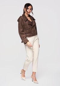 Femme portant une veste en suède marron, un pantalon crème fuselé, des chaussures à talons beige et des boucles d'oreilles dorées, posant devant un fond clair uni.