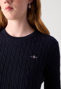 Tamsiai mėlynas kabelinis megztinis su apvaliu iškirpimu. Ypatumai: tekstūruotas raštas ir siuvinėtas GANT logotipas krūtinėje.