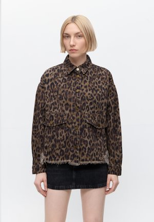 Femme aux cheveux blonds courts portant une veste marron à imprimé léopard boutonnée et une mini-jupe en denim noire, debout sur un fond blanc uni.