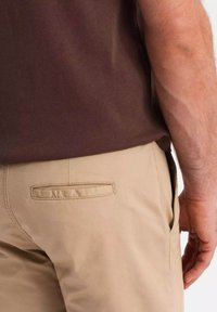 Mand iført beige bukser og brun skjorte, som viser baglommen og højre arm hængende afslappet ved siden.