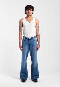 White ribbed tank top, blue wide-leg jeans, and black shoes with white accents. Model stands straight against a plain background.