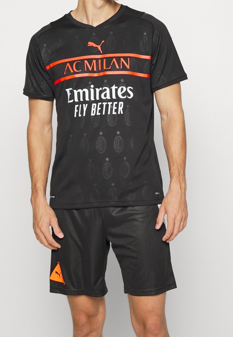 Homme portant un maillot de sport AC Milan noir avec des accents rouges et le logo "Emirates Fly Better", associé à un short noir Puma.