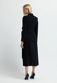 Blonde Frau in schwarzem maßgeschneidertem Blazer, plissiertem Midikleid und schwarzen High Heels steht mit dem Rücken zur einfachen Hintergrund.