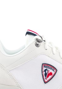 Sneaker bianco realizzato in materiali tessili e in pelle, con un'etichetta logo e un anello metallico sul lato. Accentuato da dettagli blu navy e rossi.
