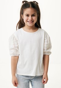 Camiseta blanca de algodón con mangas cortas abullonadas que presenta bordado tipo ojal. Textura suave, escote redondo, combinada con jeans de color azul claro.
