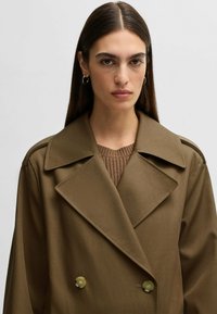 Cappotto marrone doppiopetto con ampie rever, caratterizzato da dettagli con bottoni e una texture liscia, sovrapposto a un top in maglia marrone.