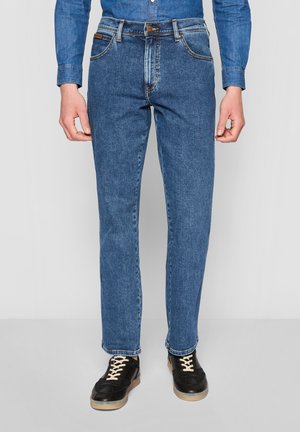 Homme portant un jean bleu slim, une chemise en denim et des baskets noires à lacets blancs, debout devant un fond gris uni.