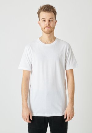 Weißes Baumwoll-T-Shirt mit Rundhalsausschnitt und kurzen Ärmeln, ausgestattet mit einem kleinen gestickten Logo auf der Brust, kombiniert mit schwarzen Hosen.