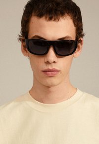 Black rectangular sunglasses with a thick frame, worn by a person in a light beige t-shirt. Sunglasses have dark lenses, minimalist design.