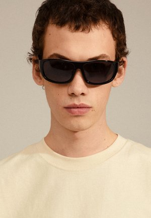 Gafas de sol negras rectangulares con un marco grueso, usadas por una persona con una camiseta de color beige claro. Las gafas tienen lentes oscuros y un diseño minimalista.