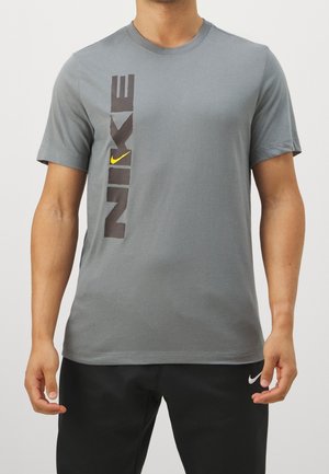 Mężczyzna w szarej koszulce Nike z pionowym czarno-żółtym logo po lewej stronie, w połączeniu z czarnymi spodniami z białym znakiem Nike.