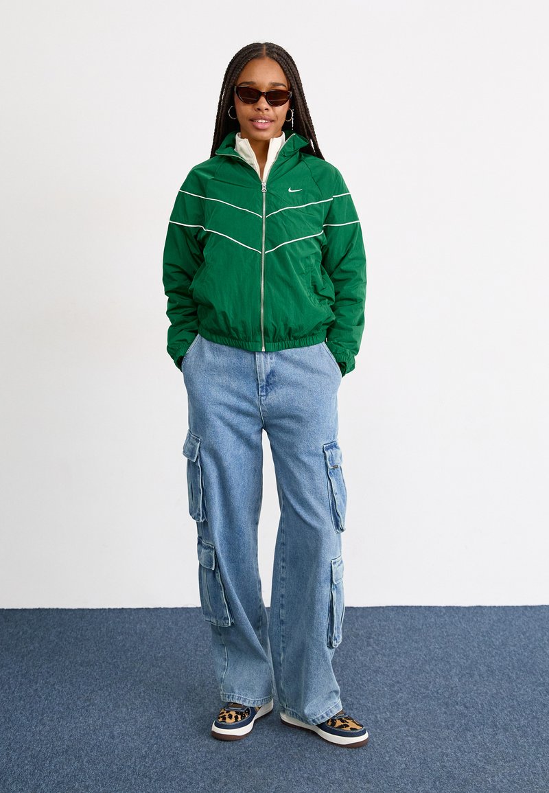 Grüne Zip-Jacke mit weißen Akzenten, hellblaue Cargo-Jeans mit Seitentaschen und braun-schwarze gemusterte Sneakers.