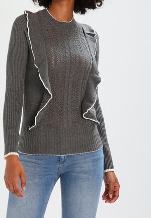 Femme portant un pull gris tricoté avec des bords à volants et une bordure blanche, associé à un jean bleu clair, sur un fond uni.
