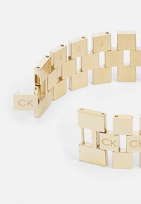 Pulsera de oro con eslabones planos y rectangulares. Cada eslabón tiene un acabado mate y presenta acentos grabados. Se cierra con un broche marcado "CK."