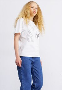 White cotton t-shirt featuring black graphic designs on the front. Paired with blue denim pants that have side pockets.