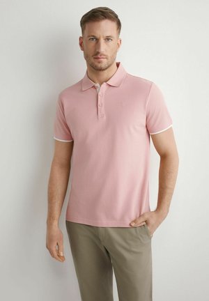 Mężczyzna z krótkimi włosami w jasnoróżowej koszulce polo i beżowych spodniach, stojący na tle jednolitego białego tła.