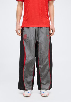 Άτομο που φοράει φαρδιά γκρι και μαύρη αθλητική φόρμα με κόκκινο περιθώριο, κόκκινη μπλούζα και λευκά αθλητικά παπούτσια με μπλε και πορτοκαλί λεπτομέρειες.