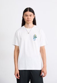 Hvid bomulds-t-shirt med et farverigt trykt grafik på venstre bryst, korte ærmer og en afslappet pasform. Bæres sammen med sorte shorts.