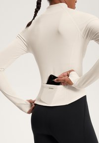 Pitkähihainen, vaaleanbeige urheilupaita, jossa on pyöreä kaula-aukko ja kädenauhat. Paidassa on takatasku puhelimelle, ja se on yhdistetty mustiin leggingseihin.