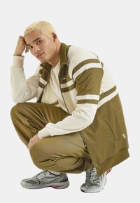 Tuta oliva e crema con motivi a righe, polsini a costine e tasche con zip; modello accovacciato in scarpe sportive grigie.