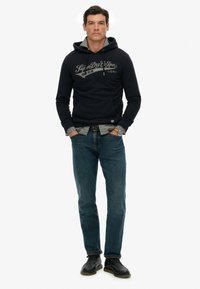 Granatowy hoodie z trokami i przednią kieszenią, szare logo, noszony na koszuli w kratę. Połączony z niebieskimi dżinsami i czarnymi butami.