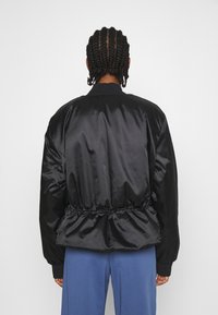 Svart satinbomberjacka med draperad fåll, ribbade ärmar och en stilren design. Bärs över blå byxor. Minimalistisk och samtida stil.