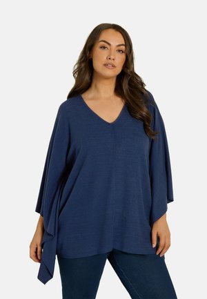 Vrouw met lang donker haar die een losse marineblauwe V-hals top en donkere broek draagt tegen een effen lichte achtergrond.