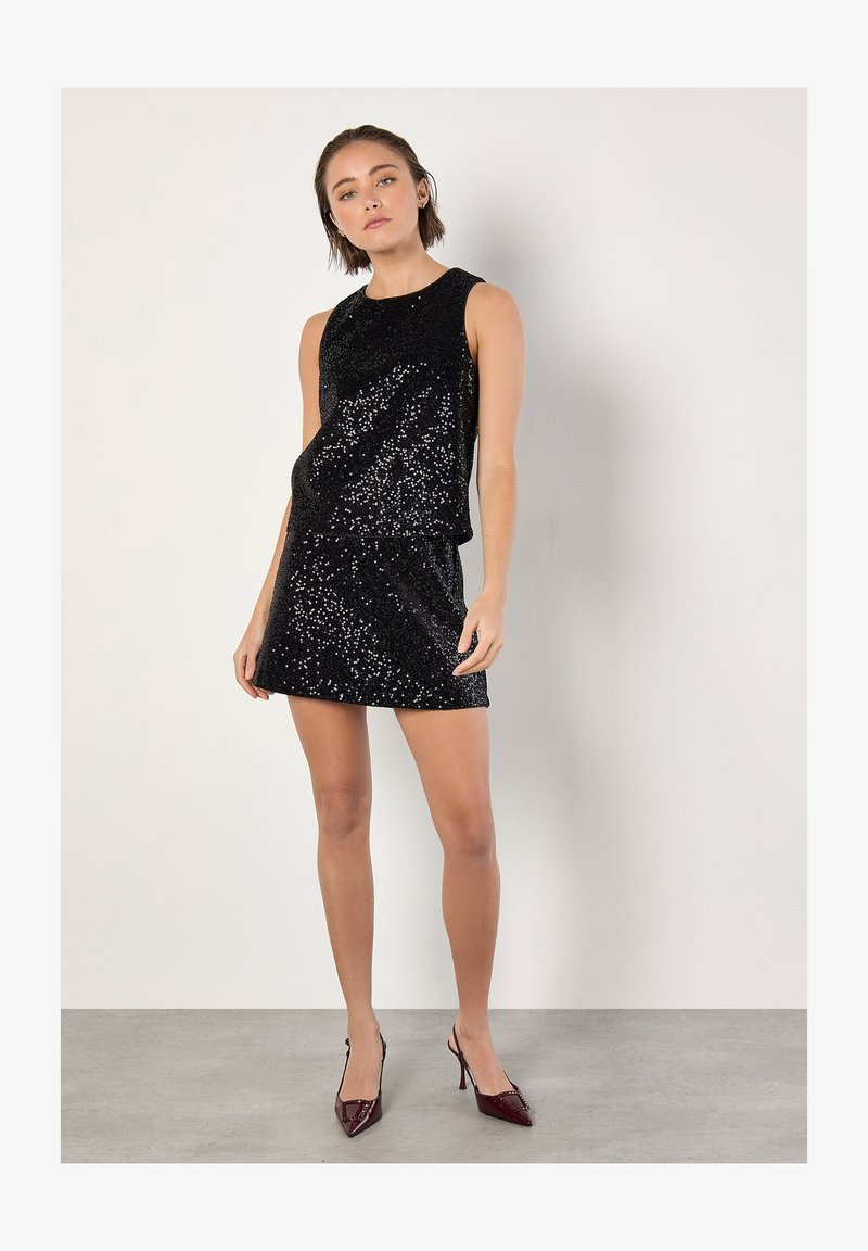 Top senza maniche in paillettes nere abbinato a una mini gonna coordinata, caratterizzata da una texture liscia e dettagli lucidi; la modella indossa scarpe con tacco a punta.