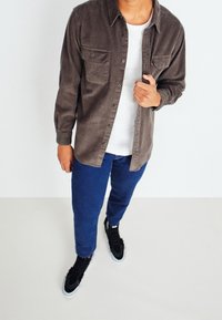 Persoon in een bruine corduroy overhemd over een wit t-shirt, blauwe jeans en zwarte sneakers staat op een lichte vloer met een witte achtergrond.
