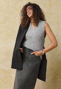 Top de punto gris sin mangas con escote en V, combinado con un abrigo largo negro y una falda de mezclilla negra de tiro alto, que incluye bolsillos.